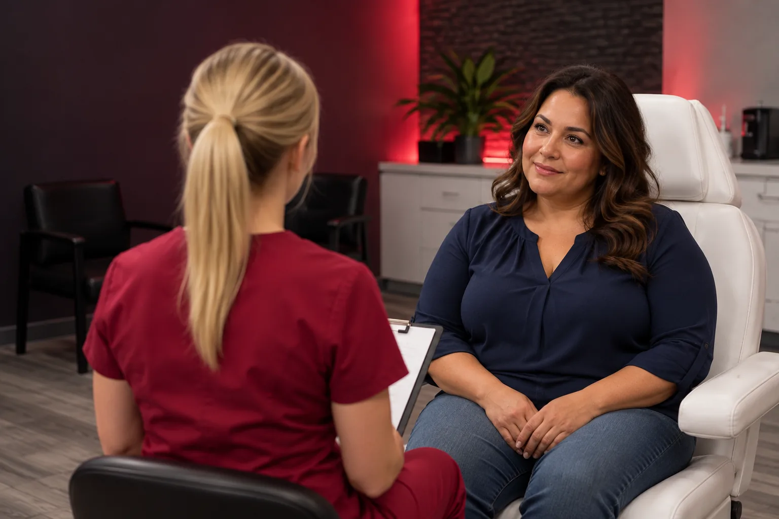 Provider in crimson scrubs with a clipboard, seen from behind, talking with a smiling woman seated in a white treatment chair
