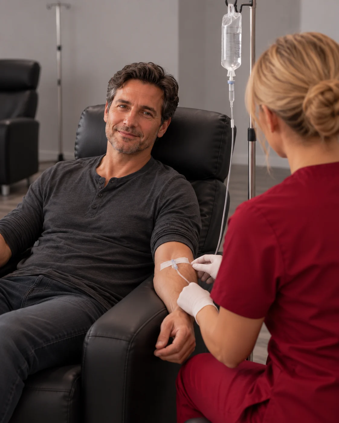 Nurse in crimson scrubs checking the IV line of a smiling man resting in a black recliner
